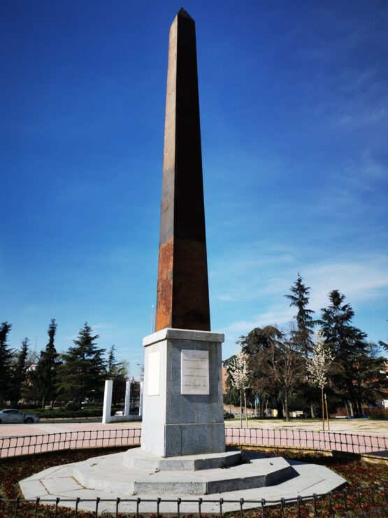 Obelisco de metal sobre un pedestal de piedra en una plaza ajardinada de Vicálvaro.